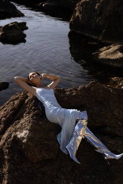 Şık gümüş elbiseli ve günbatımı kumsalında poz veren metalik yüksek çizmeli siyah saçlı güzel Slav kızın açık hava fotoğrafı.