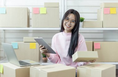 Genç Asyalı kadın dizüstü bilgisayar kullanarak ürün satın alma siparişlerini kontrol ediyor ve müşterilere teslimat için paket hazırlıyor. İnternetten ürün satıyorum. İnternetten alışveriş. İnternet işi. Küçük işletme girişimcisi KOBİ 'nin fikirlerini çevrimiçi başlatıyorum. Çalış