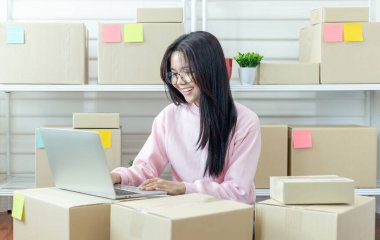 Genç Asyalı kadın dizüstü bilgisayar kullanarak ürün satın alma siparişlerini kontrol ediyor ve müşterilere teslimat için paket hazırlıyor. İnternetten ürün satıyorum. İnternetten alışveriş. Çevrim içi iş. 