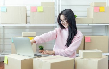 Genç Asyalı kadın dizüstü bilgisayar kullanarak ürün satın alma siparişlerini kontrol ediyor ve müşterilere teslimat için paket hazırlıyor. İnternetten ürün satıyorum. İnternetten alışveriş. Çevrim içi iş. 