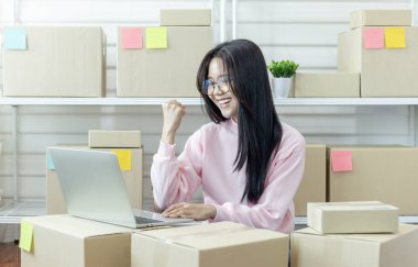 Genç Asyalı kadın dizüstü bilgisayar kullanarak ürün satın alma siparişlerini kontrol ediyor ve müşterilere teslimat için paket hazırlıyor. İnternetten ürün satıyorum. İnternetten alışveriş. Çevrim içi iş. 