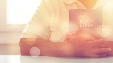 Elleri kutsal bir İncil 'in üzerinde katlanmış, ahşap bir masada dinlenen ve gün ışığıyla parlayan bir insan. Bu görüntü Hristiyan ibadetiyle inanç, bağlılık ve ruhsal bağlantıyı temsil eder..