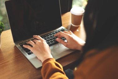 Yanında kahve fincanı olan, rahat bir kafedeki ahşap bir masada dizüstü bilgisayarda yazı yazan bir kadının yakın çekimi. Serbest çalışma kavramı, uzak iş, dijital yaşam tarzı ve modern teknoloji kullanımı.