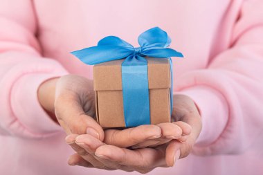 Mavi kurdeleli küçük kahverengi bir hediye kutusuyla el ele tutuşmak hediye vermeyi, kutlamayı, sürprizi ya da takdiri sembolize eder. Beyaz arka planda, pembe kazak giyen biri..