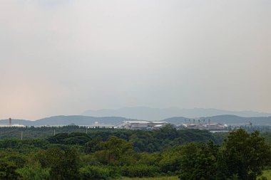 Yeşil ormanlarla çevrili endüstriyel fabrika kompleksi bulutlu gökyüzü altında uzak dağlar doğa ve endüstrinin zıtlığını gösteriyor..