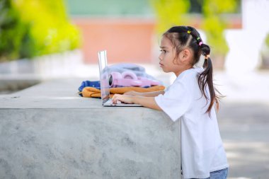 Küçük Asyalı kız okulda öğrenmek için dizüstü bilgisayar kullanıyor. Çocuk mutlu bir şekilde gülümser ve uzaktan bilgi alır. Bu da sınıfın öğrenme becerisini arttırır. Okula Dönüş Konsepti.