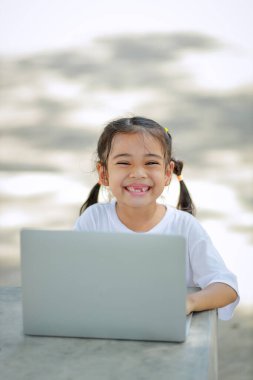 Küçük Asyalı kız okulda öğrenmek için dizüstü bilgisayar kullanıyor. Çocuk mutlu bir şekilde gülümser ve uzaktan bilgi alır. Bu da sınıfın öğrenme becerisini arttırır. Okula Dönüş Konsepti.