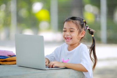 Küçük Asyalı kız okulda öğrenmek için dizüstü bilgisayar kullanıyor. Çocuk mutlu bir şekilde gülümser ve uzaktan bilgi alır. Bu da sınıfın öğrenme becerisini arttırır. Okula Dönüş Konsepti.