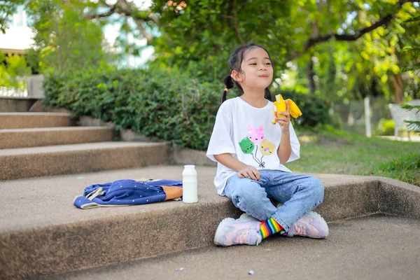 stock image Little kid girl asian holding and eat banana in the school. feel happy and enjoy eating banana  before entering the classroom.