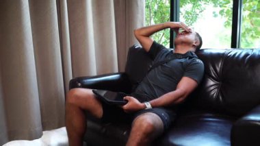 Tabletli, yorgun ve üzgün Asyalı adam evdeki kanepede oturuyor.