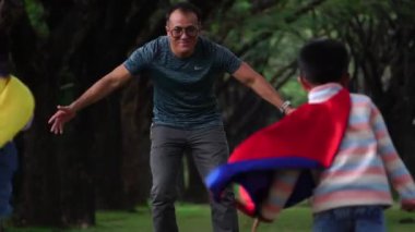 Asyalı baba parkta süper kahraman kostümleri giyip çocuklarıyla oynuyor.