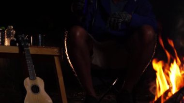  Geceleri ormanda kamp ateşinin yanında ukulele oynayan bir adam. Seyahat ve eğlence. 