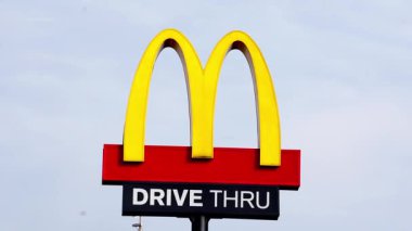 Bir McD (McDonalds) logosu, McD restoranındaki gökyüzü arkaplanlı sürücü kutusuyla birlikte ışık kutusu direğinde görüntülenir.