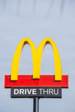 Bir McD (McDonalds) logosu, McD restoranındaki gökyüzü arkaplanlı sürücü kutusuyla birlikte ışık kutusu direğinde görüntülenir.