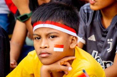 Bronzlaşmış tenli bir çocuk kırmızı ve beyaz bir saç bandı takıyor ve yanağında Endonezya bayrağı var..