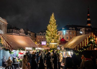Akşamları Rigas Dome Meydanı 'nda ışıltılı ışıklar ve tatil süslemeleriyle çevrili şenlikli bir Noel pazarı. Piyasa etkinliklerle dolu, ziyaretçiler mevsimlik ziyafetlerin ve el yapımı malların keyfini çıkarırken davetkar atmosfer mevcut.
