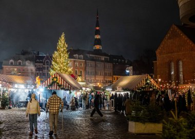 Akşamları Rigas Dome Meydanı 'nda ışıltılı ışıklar ve tatil süslemeleriyle çevrili şenlikli bir Noel pazarı. Piyasa etkinliklerle dolu, ziyaretçiler mevsimlik ziyafetlerin ve el yapımı malların keyfini çıkarırken davetkar atmosfer mevcut.