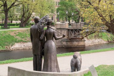 Riga şehir parkında bir adam, şemsiyeli bir kadın ve küçük bir köpeği resmeden bronz bir heykel. Rakamlar birlikte yürüyor gibi görünüyor, huzurlu bir şehir ortamında sakin ve nostaljik bir anı yakalıyorlar.