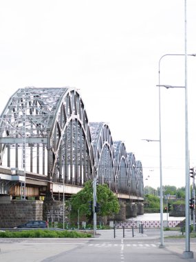 Riga, Letonya 'daki ikonik demiryolu köprüsünün Daugava Nehri boyunca uzanan ayırt edici metal kemerleriyle manzarası. Kompozisyon, kentin kentsel zeminine karşı kurulan köprünün endüstriyel mimarisini ve simetrisini vurguluyor.. 