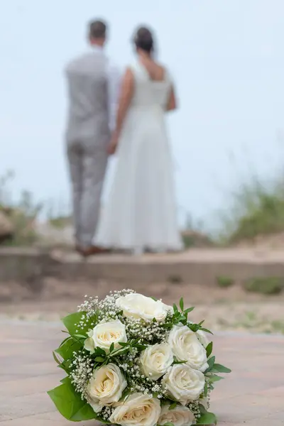Beyaz güllü bir gelin buketi ön planda zarafet ve saflığı sembolize ederek hafifçe yerde yatar. Arka planda, bir gelin ve damat yan yana durur, mesafeye bakarlar..