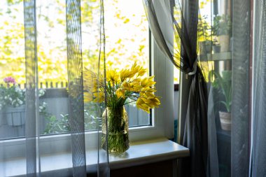 Sıcak doğal ışıkla hafifçe aydınlatılan sarı lalelerle dolu bir vazo. Balkon kapısı hafifçe açık, hafif bir esinti ve sakinlik ve açıklık hissi katıyor. Bahar atmosferiyle dolu sıcak, güneşli bir an..