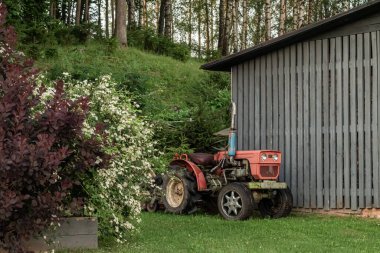 Kırsal bir traktör, yemyeşil çimenler ve canlı açan kır çiçekleriyle çevrili geleneksel bir ahırın yanında duruyor. Kırsal bir çiftliğin huzurlu ve zamansız atmosferini yakalıyor..