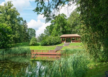 Durgun suyun üzerinde uzanan küçük bir göleti ve ahşap iskelesi olan huzurlu bir yaz manzarası. Yakınlarda, yeşil çimenler ve açık mavi gökyüzünün altındaki ağaçlarla çevrili, dinlenmek ve dinlenmek için mükemmel, rahat, küçük bir kabin bulunur..