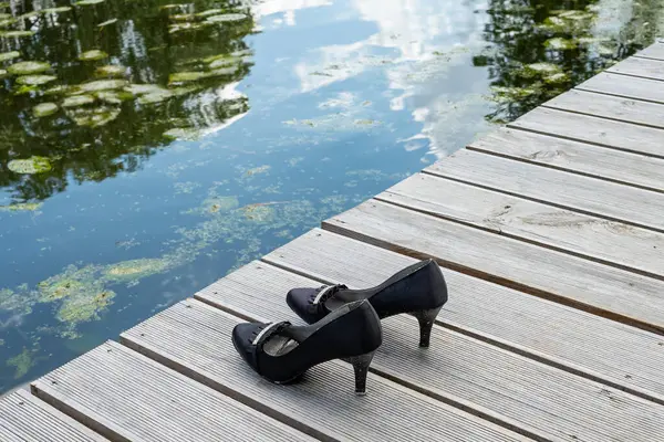Bir çift siyah yüksek topuklu ayakkabı, sakin bir göletin yanındaki eski ahşap tahtalara dikkatlice yerleştirilmiş. Sakin su, sakin çevreyi yansıtır, zarif ayakkabılar ve doğal, kırsal ortam arasında bir kontrastlık yaratır.. 