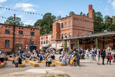 Eski bir kırmızı tuğlalı endüstriyel komplekste kutlama. İnsanlar kiraz kuşları ve dekorasyonların ortasında eğleniyorlar, tarihi mimariyi neşeli sosyal aktivitelerle harmanlıyorlar, canlı ve renkli bir sahne yaratıyorlar..
