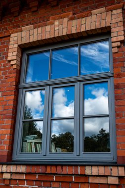 Kırmızı tuğlalı bir binada büyük bir pencere. Pencere çerçeveyi doldurur, camdaki bulutları yansıtır, kitaplar pencere kenarına yerleştirilir, mimari detay ve şiirsel atmosfer karışımı yaratır..