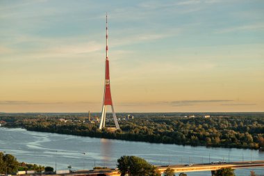 Daugava Nehri 'nin kıyısındaki televizyon kulesinin gün batımında gökyüzünü dolduran sıcak turuncu renkler. Sahne sakin ve atmosferik bir anı yakalıyor, kuleyi parlayan akşam arkaplanına vurguluyor..