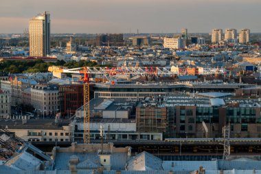 Wide panoramic view of a city with a construction site in the foreground, featuring a tall yellow crane and urban skyline in the background under natural daylight.
