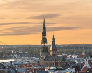Riga kilise kuleleri, sıcak altın, turuncu ve pembe renkleri ile günbatımı gökyüzüne karşı kurulmuş, tarihi gökyüzünü aydınlatan. Sahne, şehrin mimari güzelliğini ve dingin akşam atmosferini yakalıyor..