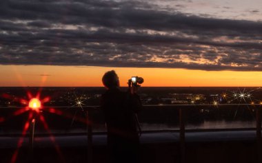 Sunset cityscape with a photographer capturing the beautiful view of the city at dusk. The warm evening light reflects on the skyline, creating a serene and atmospheric scene filled with golden and orange tones.