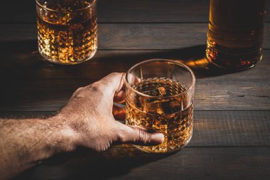 Hand holding whisky glass with ice on wooden table background. Close up.