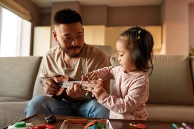 Little girl setting up ukulele in hands of her father on sofa at home. Asian man and girl spending time together. Domestic entertainment and leisure. Family relationship. Parenting and fatherhood