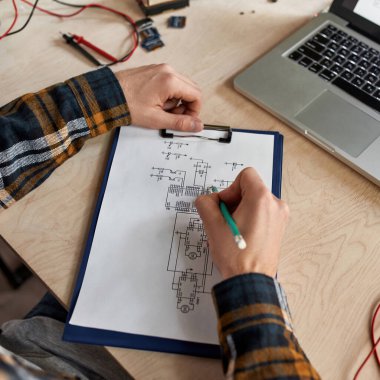 Cropped male IT technician or engineer editing technical scheme with pencil. Man at table with laptop, variety technical tools and components. Modern technology and innovation development