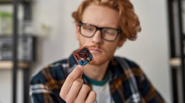 Selective focus of microchip in hand of concentrated blurred partial male IT technician or engineer looking at him. Young red haired caucasian man wearing glasses. Modern technology and innovation