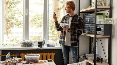 Concentrated male IT technician or engineer looking at microchip and holding plastic box in sunny office. Young red haired caucasian man wearing glasses. Modern technology and innovation development