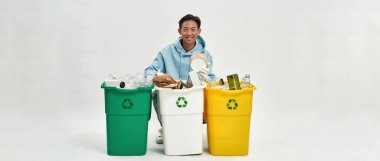 Smiling asian man sort garbage in glass, plastic and cardboard dustbins and look at camera. Ecology safety and protection. Waste disposal and recycling. White background. Studio shoot. Copy space
