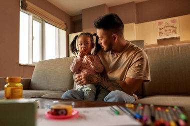 Father looking at daughter in his arms eating multivitamin jelly on sofa at home. Asian man and little girl spend time together. Family relationship. Health and medical care. Parenting and fatherhood