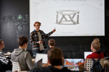 Genç beyaz erkek öğretmen, ders odasında ya da sınıfta öğrencilere robotik modelin detaylarını tahtada gösteriyor. Modern bilişim, teknik ve mühendislik eğitimi. Okul, üniversite, enstitü