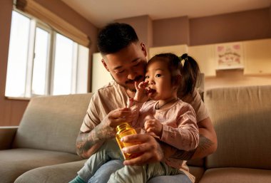 Baba, evdeki koltukta babasının kollarında otururken kızına multivitamin jölesi yerken bakıyor. Asyalı bir adam ve küçük bir kız. Aile ilişkisi. Sağlık ve tıbbi bakım. Ebeveynlik ve babalık