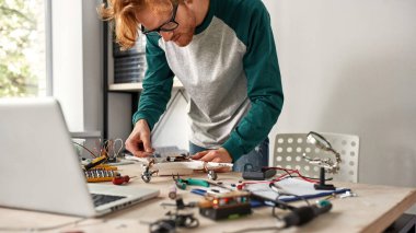 Yoğunlaşmış erkek bilişim teknisyeni ya da mühendis çeşitli teknik aletler ve parçalarla masadaki bozuk drona bakıyor. Gözlük takan genç kızıl saçlı adam. Modern teknoloji ve yenilik