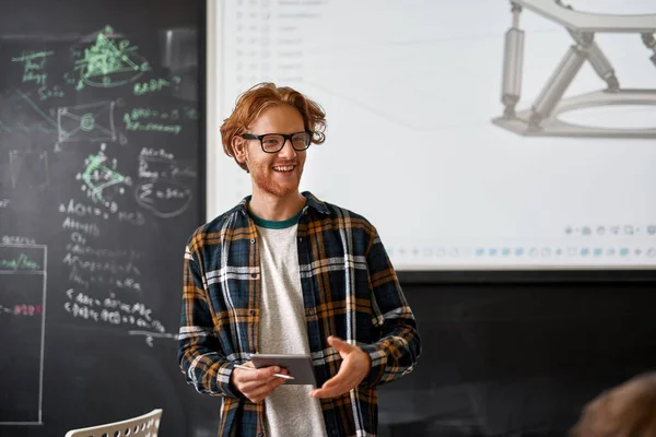 Young smiling male caucasian teacher looking at audience during teaching robotics students in classroom. Man wear glasses. Modern IT, technical and engineering education. School, college, institute