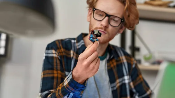 Selective focus of microchip in hand of focused partial male computer technician or engineer looking at him. Young red haired caucasian man wearing glasses. Modern technology and innovation