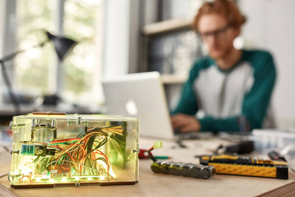 Selective focus of processor unit of robot with blurred background of male IT technician using laptop in office. Man sitting at table with tools and components. Modern technology and innovation
