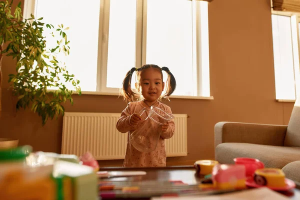 Asian little girl with glass jar looking at blurred table with toys at home. Cute female child wear pajama. Domestic entertainment and leisure. Modern childhood lifestyle. Spacious flat at sunny day