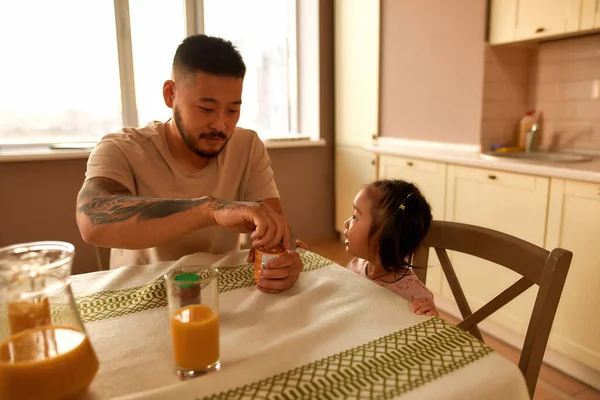 Asian little girl looking at father opening jar with baby food for her at table on home kitchen. Family relationship. Parenting and fatherhood. Healthy eating. Domestic lifestyle. Sunny day