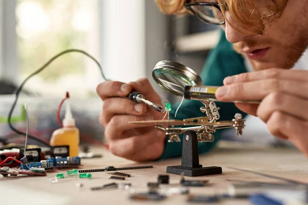 Cropped male engineer wearing glasses soldering microchip with soldering iron under magnifying glass at table with blurred variety technical tools and components. Modern technology and innovation